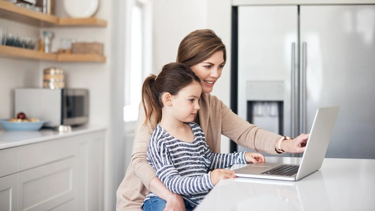 A mother and her young daughter sitting in a modern kitchen, looking at a laptop computer