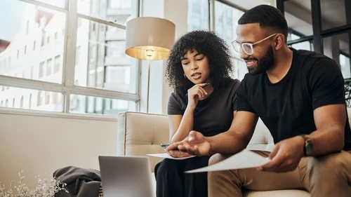 Man and woman sitting on a sofa, looking at a computer screen and discussing paperwork